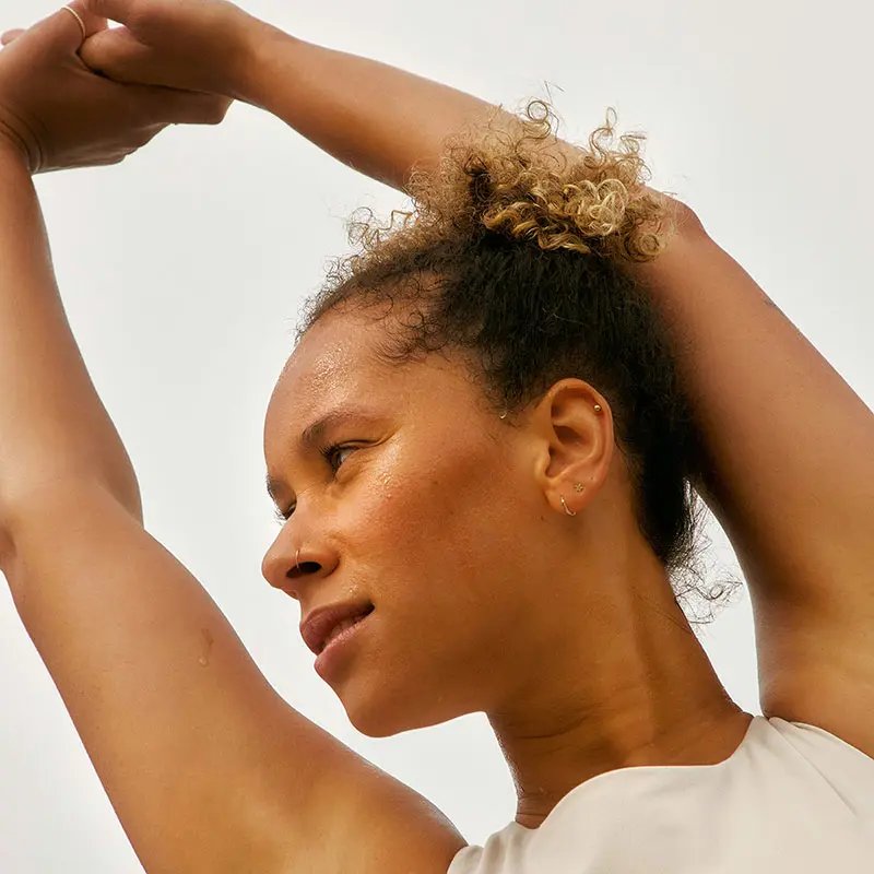 A young woman stretches her arms high above her head, gazing upwards. She has curly hair tied back and glistening skin, suggesting an active lifestyle.
