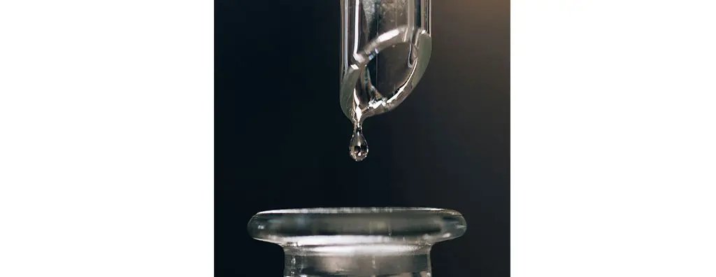 A clear drop of liquid is about to fall from a glass pipette into a round glass container below, set against a blurred dark background, creating a serene atmosphere.