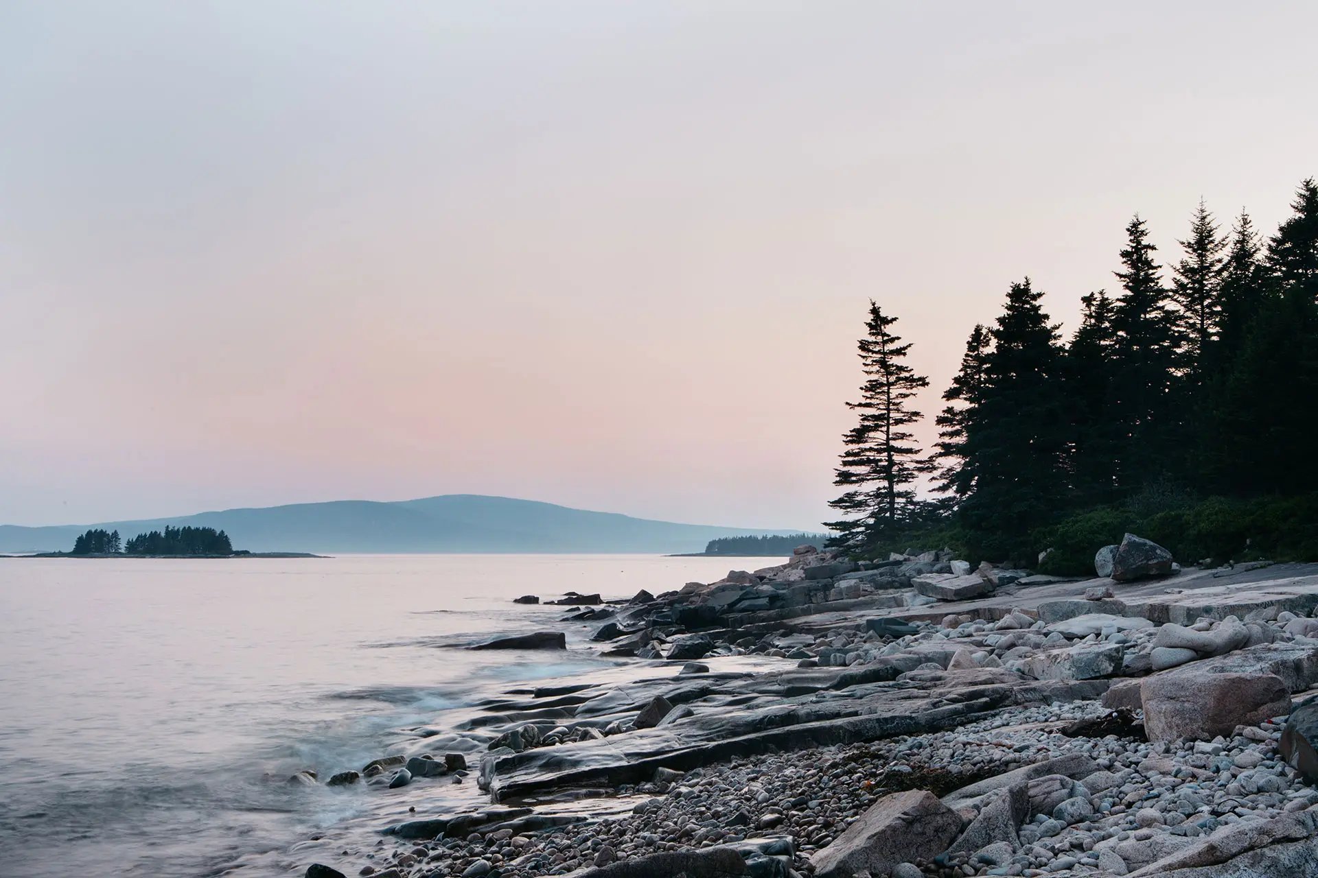 A serene coastal scene at dusk, featuring smooth stones and rocky shores with tall evergreen trees stand on the right.