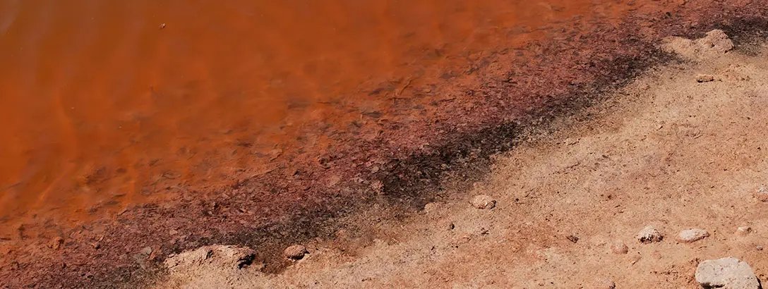 A shallow shoreline where vibrant orange water meets a dusty brown bank, showing contrasting textures.