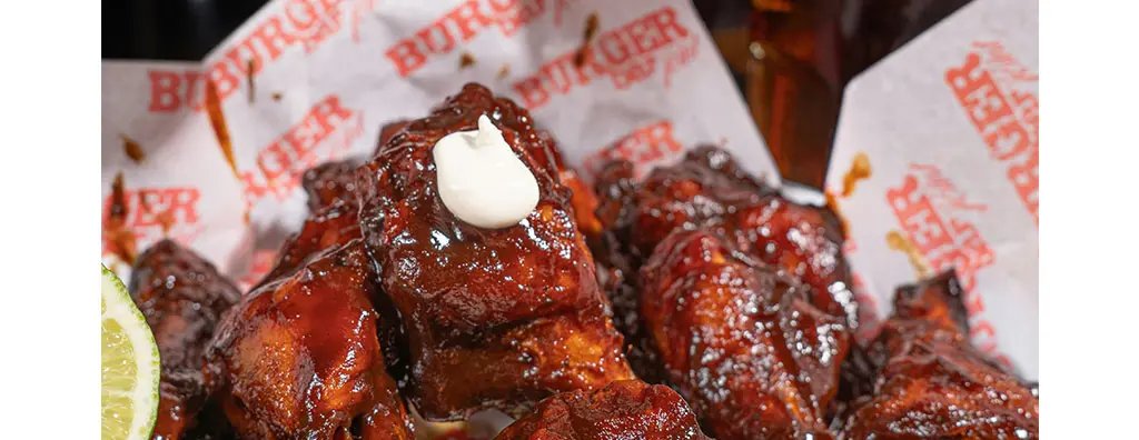 A close-up of glazed chicken wings topped with a dollop of creamy sauce, arranged in a basket lined with branded paper.