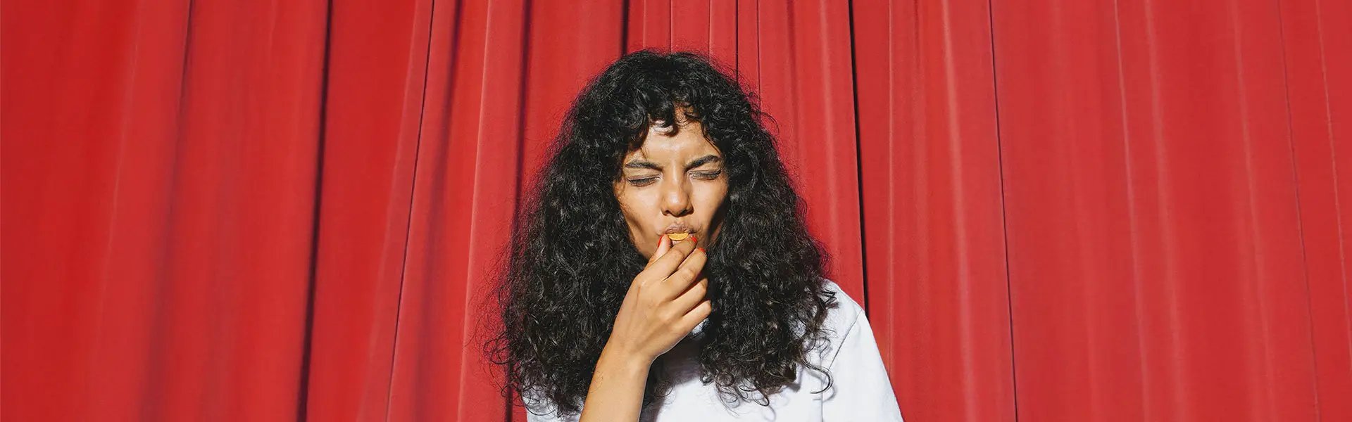 A person with curly hair is tasting some very spicy food while standing in front of a vibrant red curtain.