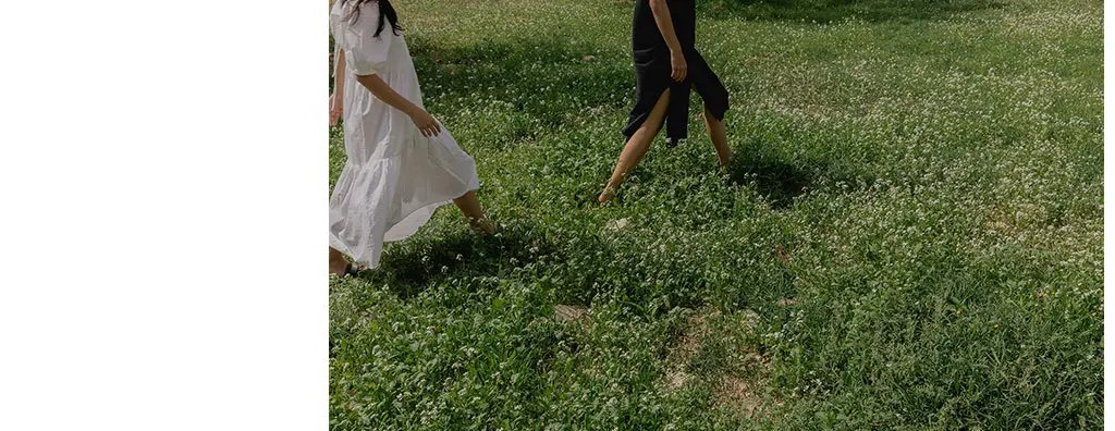 Two individuals walk through a lush green meadow filled with white flowering plants. One wears a flowing white dress and the other a black outfit.