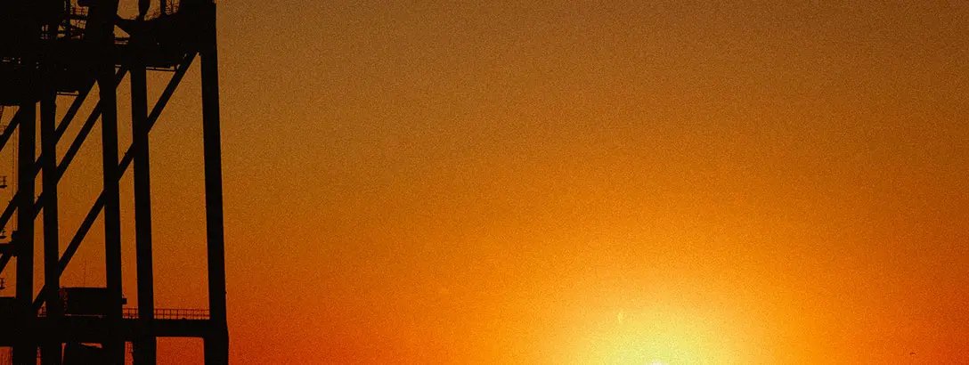 A vibrant sunset casts warm orange and yellow hues across the sky and shimmering water. In silhouette, industrial structures frame the scene, while a distant skyline adds depth.