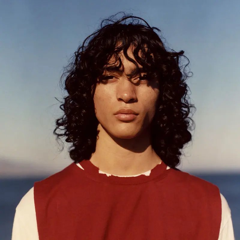 A young person with long, curly hair stands against a clear blue sky near the water, wearing a red sleeveless top over a white shirt. 