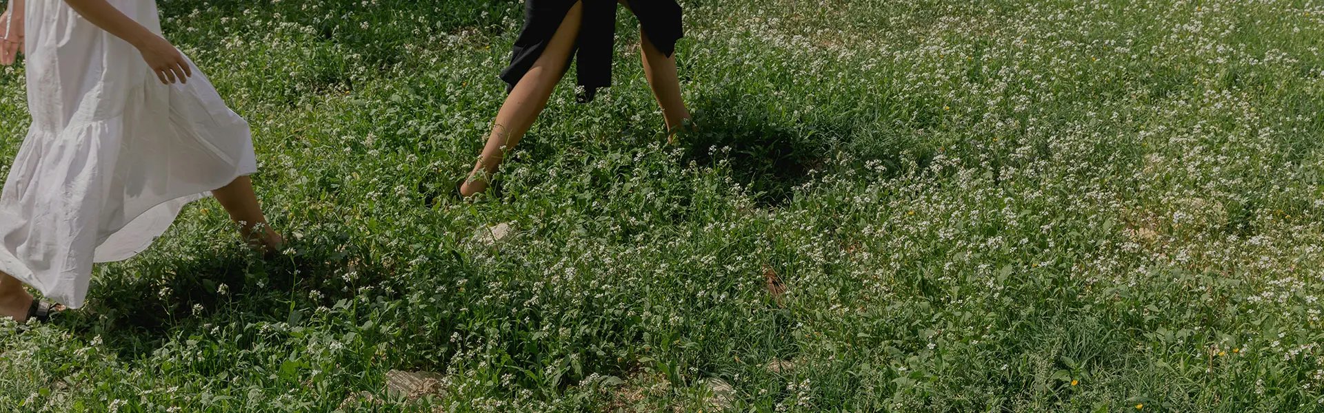 Two individuals walk through a lush green meadow filled with white flowering plants. One wears a flowing white dress and the other a black outfit.