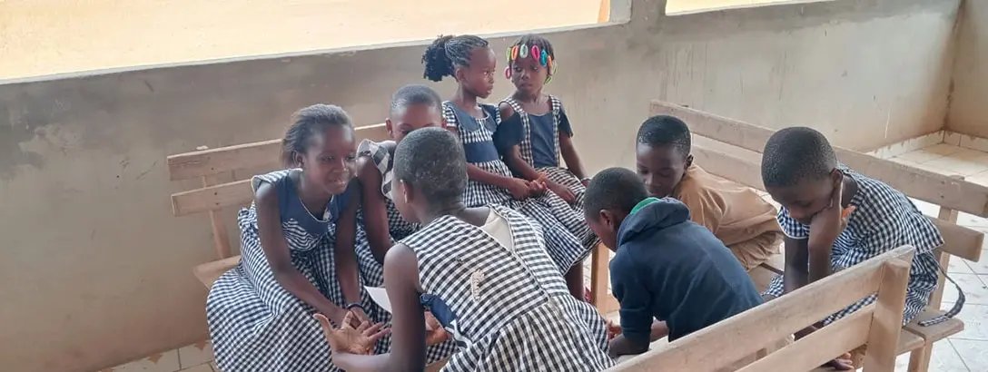 A group of children in checkered dresses are seated on wooden benches in a classroom. They are engaged in a discussion, with some interacting closely, while others observe thoughtfully.