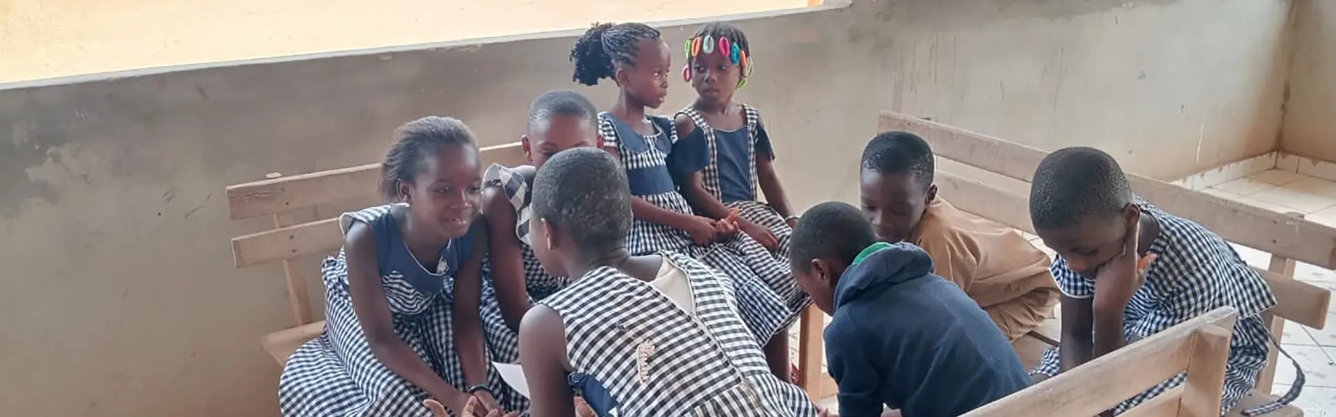 A group of children in matching checkered dresses gather on wooden benches, engaged in a lively conversation. The setting is bright and open, with a sandy area visible outside.