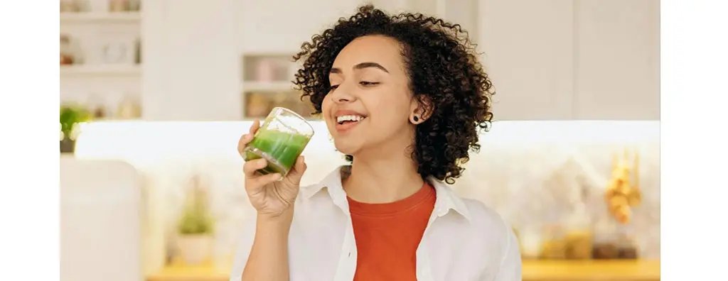 A person with curly hair smiles while holding a glass of green drink in a bright kitchen. The background features shelves with plants and cooking jars, creating a vibrant atmosphere.