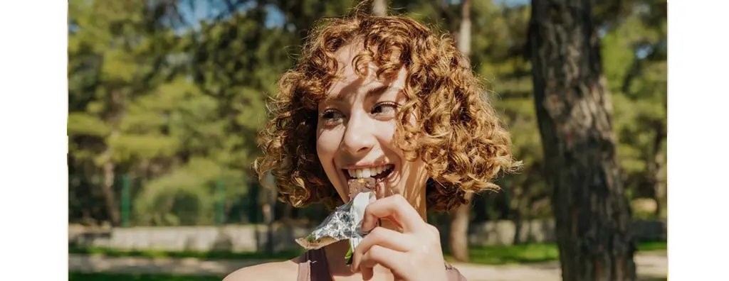A cheerful person with curly hair enjoys a chocolate bar, smiling brightly in a sunlit outdoor setting surrounded by trees. The mood is joyful and carefree.