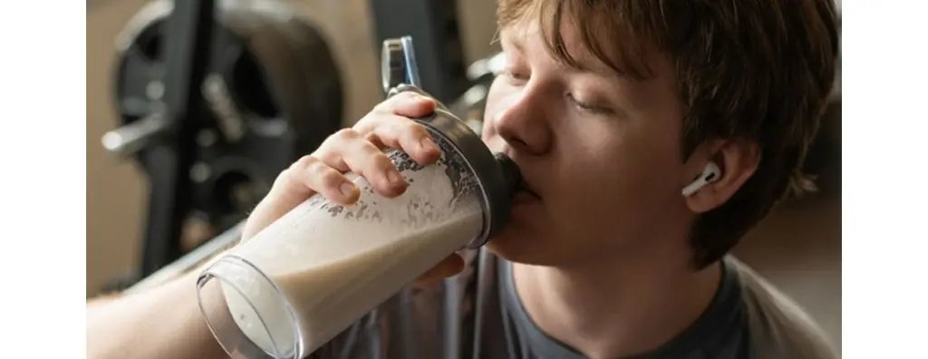 A young person with light brown hair takes a sip from a transparent shaker bottle filled with a creamy drink. They wear white wireless earbuds, and a blurred gym setting is visible in the background, evoking a sense of post-workout enjoyment.