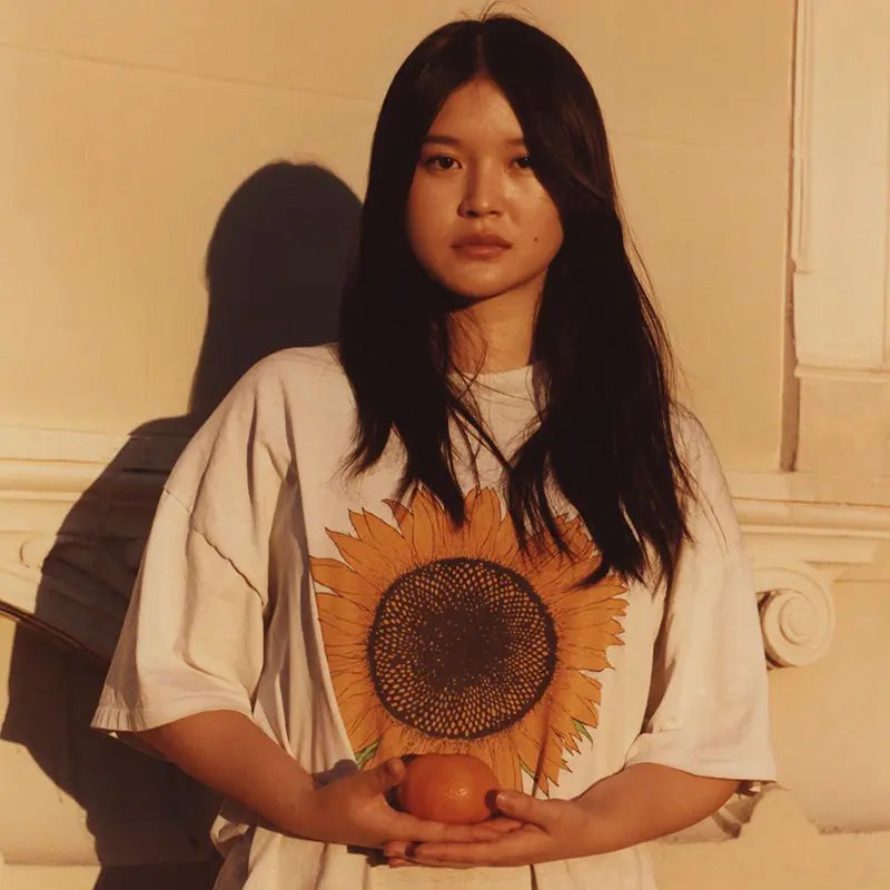 A young woman stands against a light-coloured wall, wearing a white T-shirt featuring a large sunflower graphic. She holds an orange in her hands, with soft, warm lighting creating a serene mood.