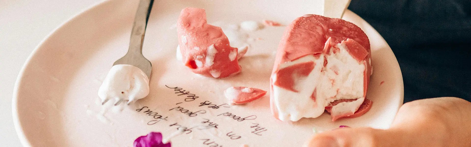 A hand holds a white plate with pink and white ice cream pieces, a silver fork, and purple flower petals scattered. The plate features a handwritten message, creating a playful and indulgent mood.