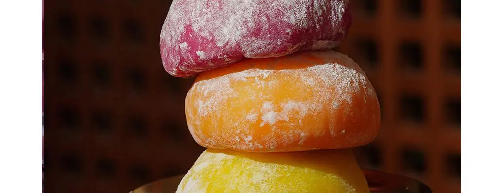 Three vibrant mochi pieces are stacked: a purple one on top, an orange one in the middle, and a yellow one at the bottom, all lightly dusted with powdered sugar. The background features a warm, woven texture, creating a cosy atmosphere.
