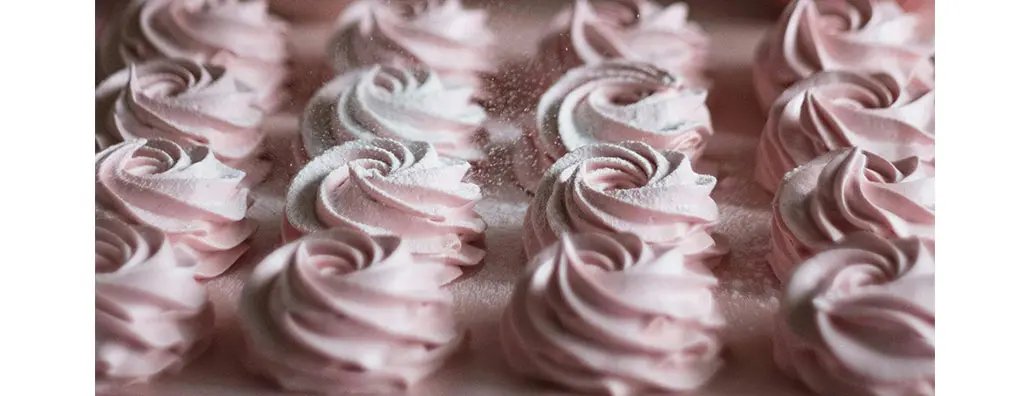 A close-up view of delicate pink swirled meringues arranged in neat rows. Each meringue is dusted with a light coating of icing sugar, creating an elegant and inviting appearance.