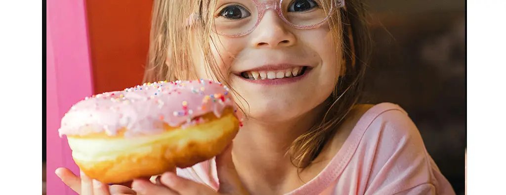 A cheerful child with brown hair wears pink glasses and smiles widely while holding a large pink frosted doughnut covered in colourful sprinkles. The background features soft, warm lighting with hints of pink, creating a playful and joyful atmosphere.