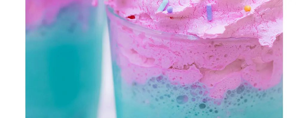 Swirls of pastel-coloured icing in soft pink, blue, and white top off a beverage generously scattered with vibrant confetti sprinkles, creating a joyful and playful treat.