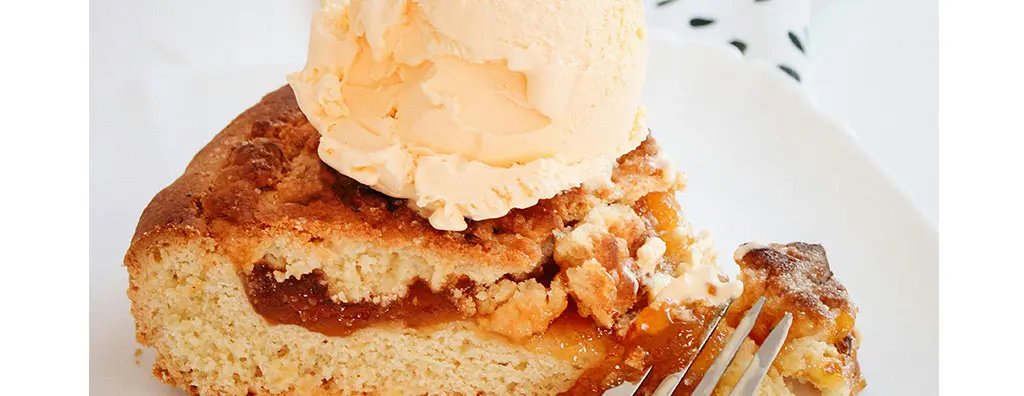 A slice of moist, golden cake topped with a scoop of creamy orange ice cream sits on a white plate, alongside a silver fork. The dessert looks inviting and indulgent.