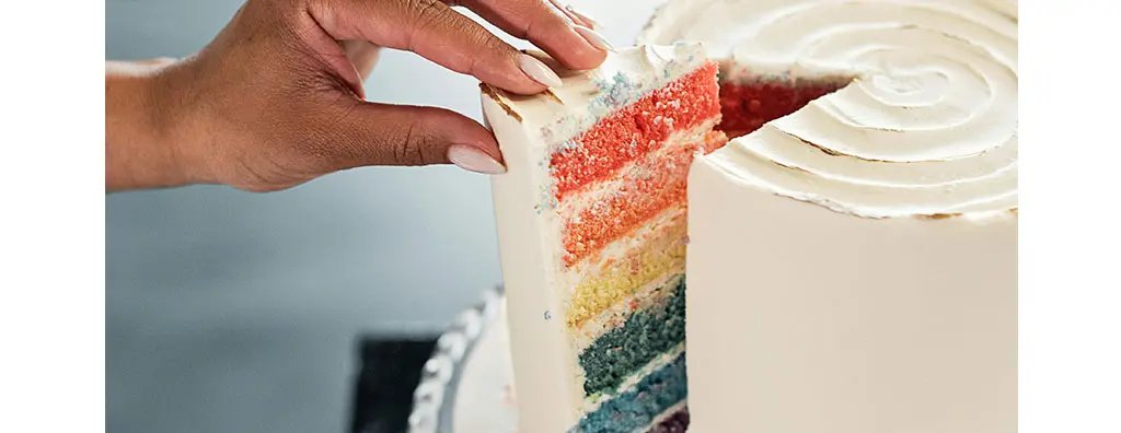 A hand is lifting a slice from a visually striking layered cake, showcasing vibrant layers of red, orange, yellow, and blue cake beneath a smooth white icing. The setting has a soft background, evoking a cheerful and inviting mood.