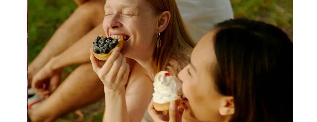 Two women enjoy sweet desserts outdoors, laughing and smiling. One holds a cupcake topped with blueberries, while the other has a cupcake with whipped cream and a cherry, set against a green background.