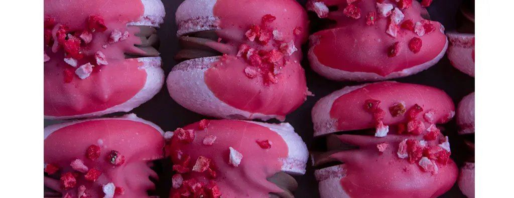 Delicious pink desserts with a glossy chocolate filling, topped with crushed freeze-dried strawberries. The sweets are arranged neatly against a dark background, enhancing their vibrant colours.