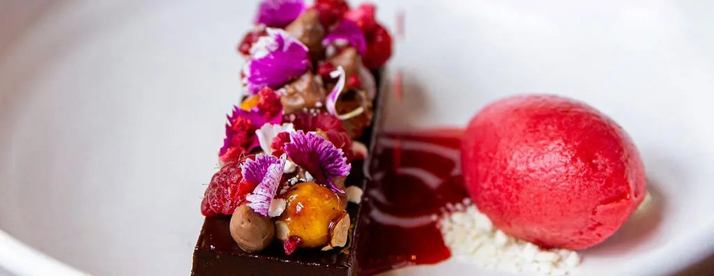 A refined chocolate dessert is elegantly plated with bright edible flowers and raspberries, accompanied by a vibrant raspberry sorbet and a drizzle of sauce.