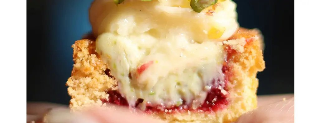 A close-up of a dessert featuring creamy filling topped with chopped pistachios and a slice of strawberry, resting on a golden-brown pastry base, with a blurred background.