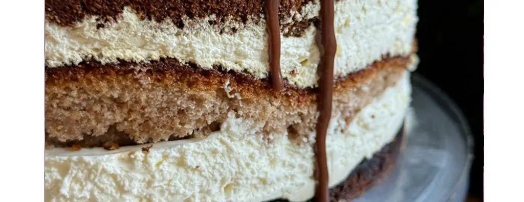 A close-up view of a layered cake showcasing soft sponge layers separated by light, fluffy cream. A drizzle of chocolate sauce runs down the side, enhancing the indulgent appearance.