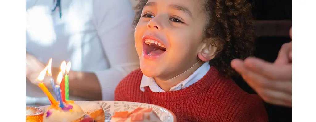 A joyful child with curly hair smiles widely, celebrating with a plate of colourful cupcakes and candles. The warm lighting enhances the cheerful atmosphere of the occasion.
