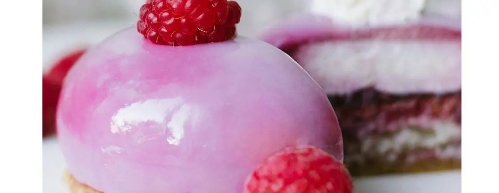 A glossy, pink dessert topped with a fresh raspberry sits in focus, with a second, layered cake partially visible behind. The scene has a light, indulgent feel.