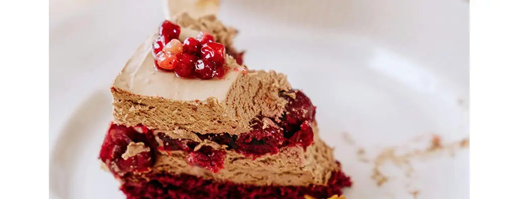 A slice of rich chocolate mousse cake sits on a white plate, topped with glossy red berries. The layered texture reveals a chocolate filling interspersed with fruit, evoking a feeling of indulgence.