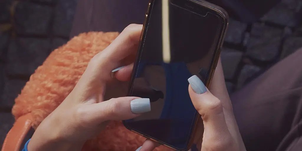 A pair of hands with light blue nails holds a smartphone, its screen dark and reflecting light. The background features a textured surface, suggesting an outdoor setting.