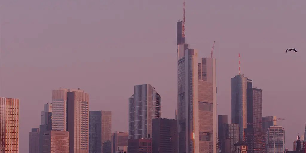 A city skyline at twilight featuring tall, modern skyscrapers with reflective glass. The sky is tinted pink and purple, creating a serene atmosphere. A bird is seen flying in the foreground.