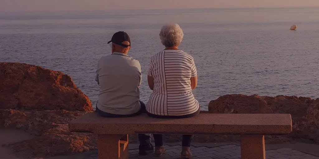 A couple sits closely together on a stone bench, looking out over the calm sea at sunset. The sky is softly lit with warm colours, creating a tranquil atmosphere.
