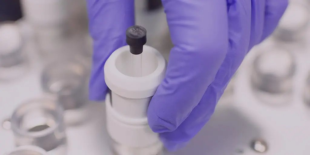A gloved hand holds a white cylindrical component with a black top, possibly part of scientific equipment, against a blurred background of metal parts. The setting suggests a laboratory or technical workspace.