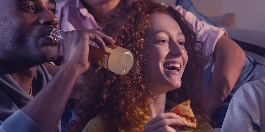 A group of friends enjoys a relaxed evening together, with a woman laughing and holding a slice of pizza, while a man drinks from a bottle. The warm lighting creates a cheerful atmosphere.