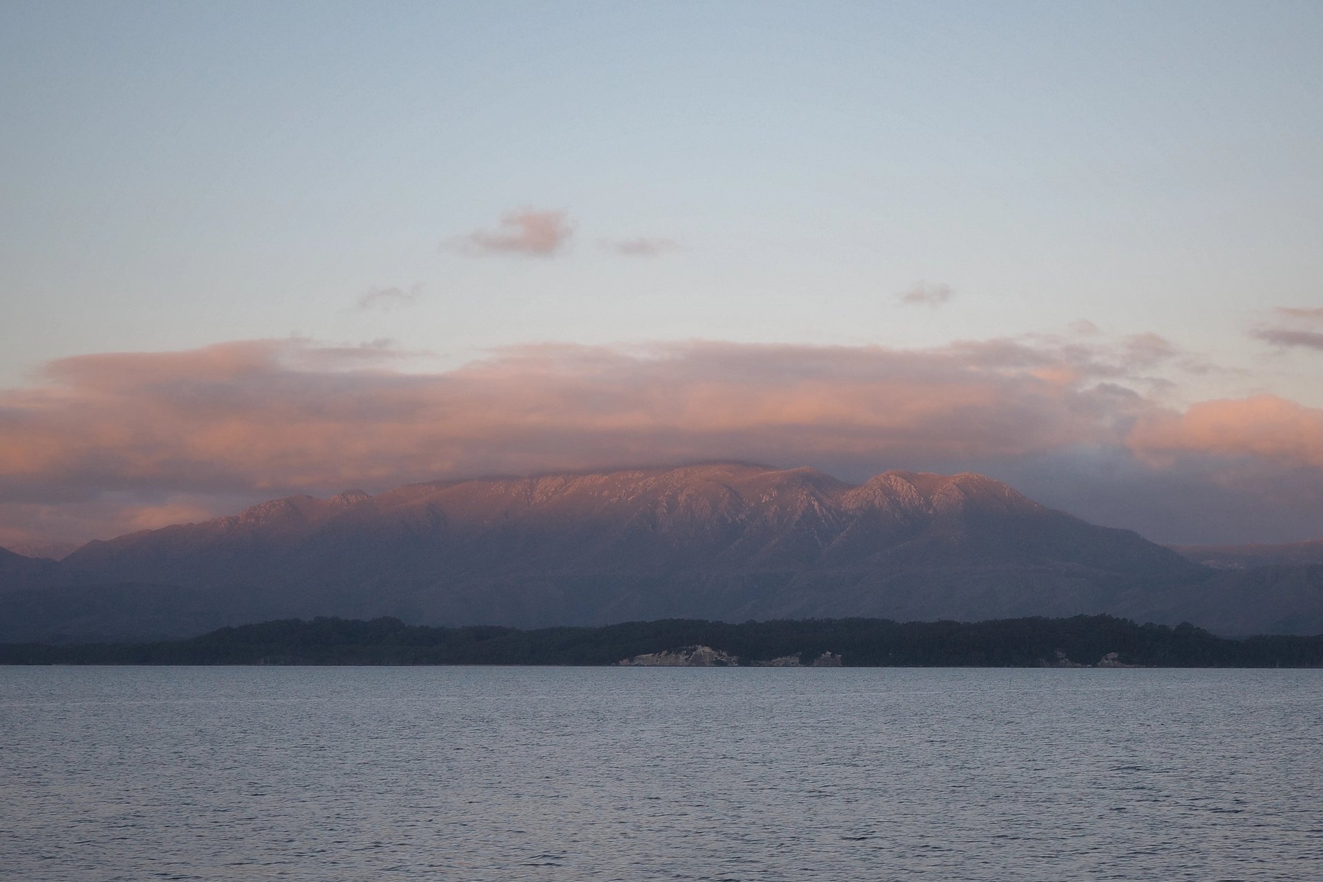 The image showcases a distant mountain range under a soft blue sky, with early evening clouds tinted in pink and orange hues. The calm water in the foreground reflects the serene landscape, creating a tranquil atmosphere.