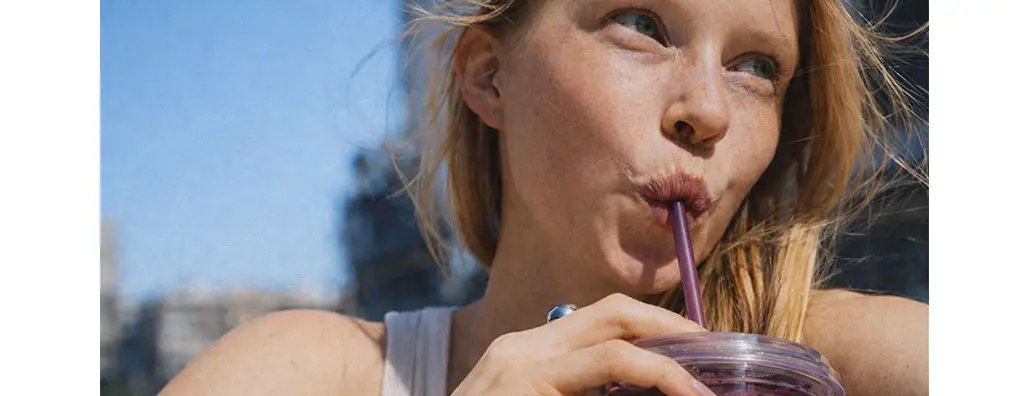 A woman with long, flowing hair enjoys a purple smoothie outside on a sunny day, sipping through a purple straw. She has a relaxed expression and wears a light tank top.