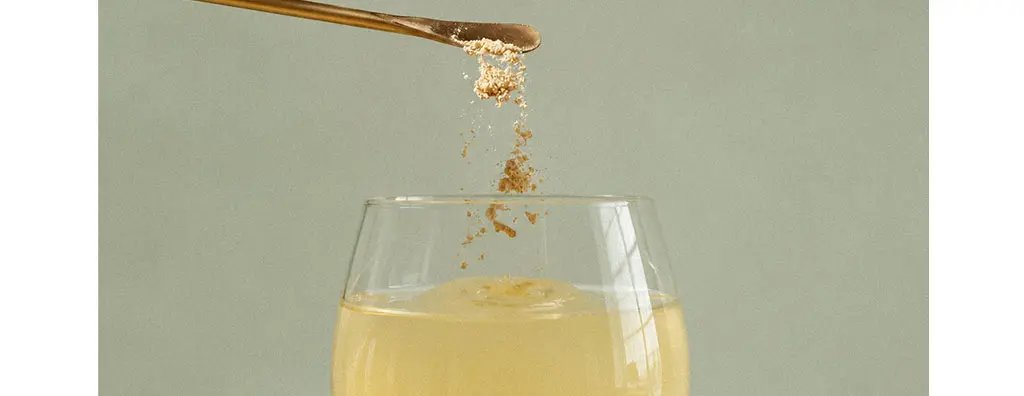 A golden liquid swirls in a clear glass, with a gold spoon pouring fine powder over the surface. The soft green background enhances the calm, sophisticated feel of the scene.
