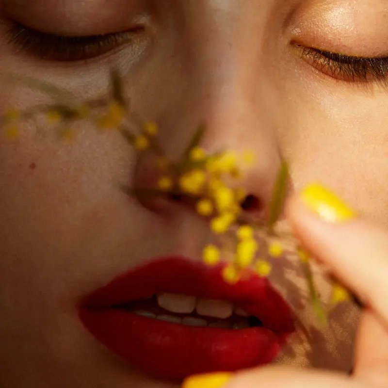 A close-up of a person's face with striking red lips and golden-yellow nail polish, gently touching a small flowering branch. The eyes are closed, conveying a serene mood.