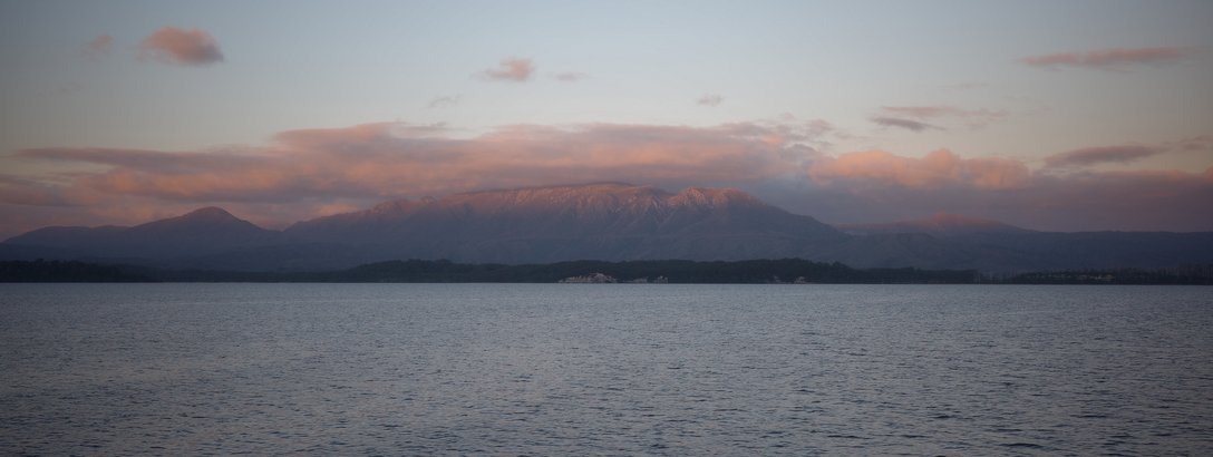 Calm water with a distant shoreline and a mountain range partially covered by clouds at sunset.