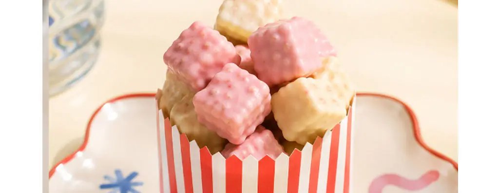 A small striped red and white box filled with pastel-coloured, square-shaped snacks sits on a decorative plate.