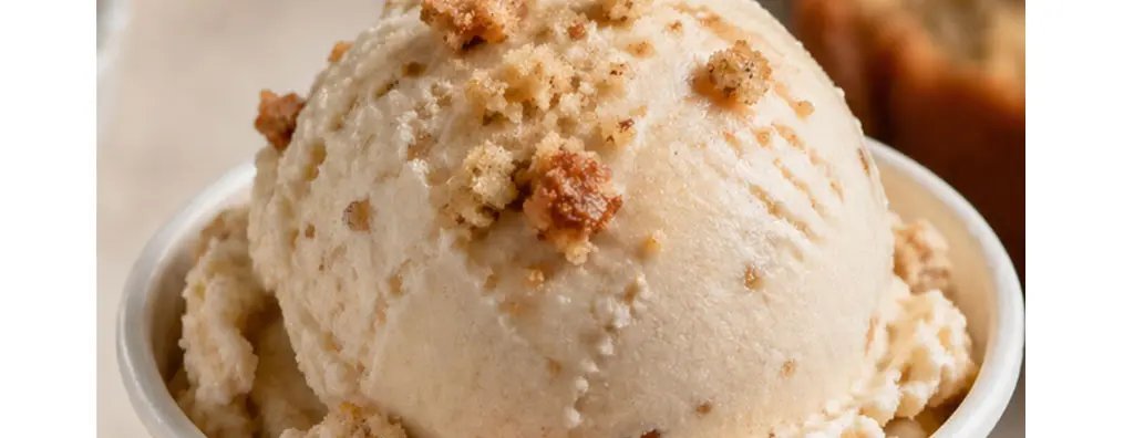 A creamy scoop of ice cream sits in a plain, beige cup, topped with small brown crumbs. The setting is soft and warm, evoking a comforting dessert vibe.