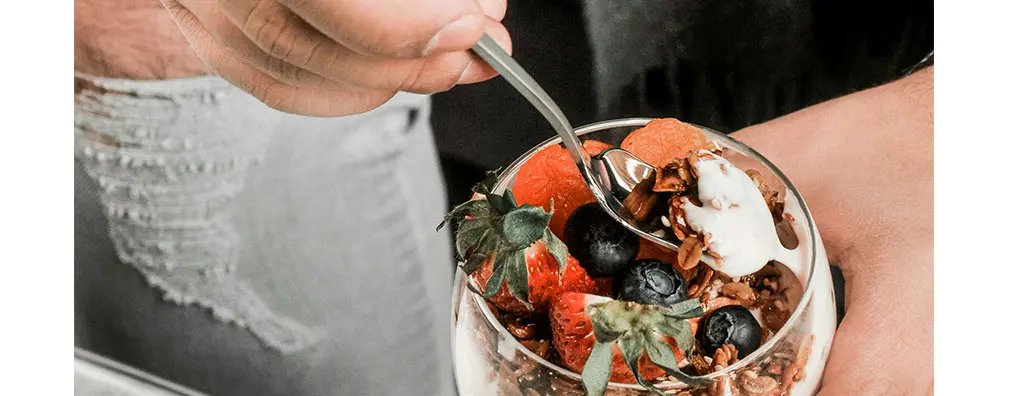 A hand holds a silver spoon, poised above a glass dessert filled with layers of yogurt, granola, strawberries, and blueberries, set on a wooden surface. 