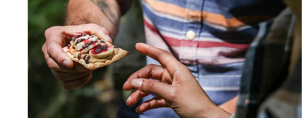 A person in a striped shirt offers a crispbread topped with spreads and toppings, while another individual reaches out to take it.