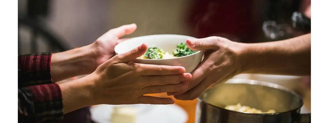 Two pairs of hands reach out to exchange a steaming bowl of pasta topped with broccoli, set against a softly blurred indoor background. 