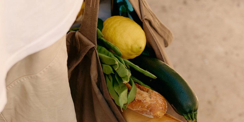 Close-up of a person carrying a brown reusable grocery bag filled with a lemon, leafy greens, zucchini, and a crusty loaf of bread.