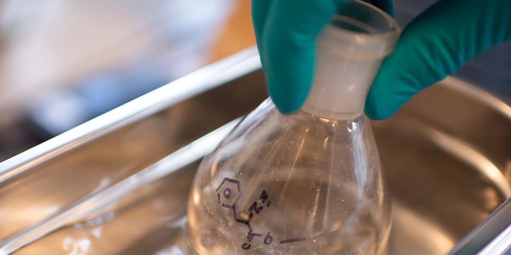 Gloved laboratory hands hold a labeled glass Erlenmeyer flask in a metal tray, highlighting careful scientific sample handling.