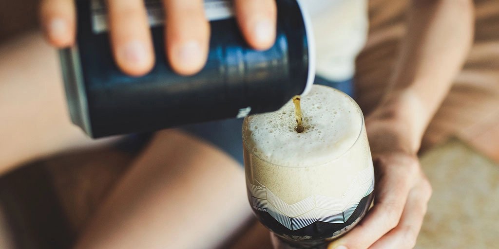 Hands pour beer from a black can into a clear glass, creating a thick foamy head; the warm scene conveys a casual, refreshing mood