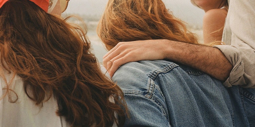 Close-up of friends relaxing outdoors, with a person’s arm resting gently on a companion in blue jeans; warm sunlight creates an intimate, carefree mood.